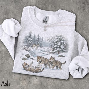 May include: A light grey heather crewneck sweatshirt featuring a graphic print of three wolf pups playing in a snowy forest landscape with pine trees and a stream. The garment has long sleeves with ribbed cuffs and a Gildan brand label at the neck.