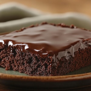 Puede incluir: Una porción cuadrada de pastel de chocolate con un glaseado brillante de chocolate negro. El pastel está sobre un plato verde y marrón. La imagen muestra un primer plano del pastel, ideal para un postre dulce.
