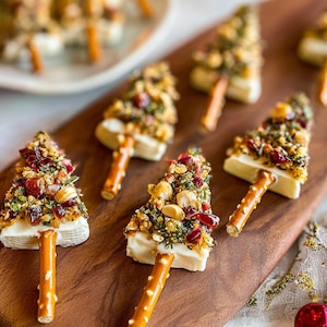 Puede incluir: Aperitivos con forma de árbol de Navidad sobre una tabla de madera. Cada árbol está hecho con una base blanca, cubierta con una mezcla de nueces, hierbas y arándanos rojos secos, y tiene un tronco de palito de pretzel.
