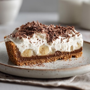 Puede incluir: Una porción de tarta de crema de plátano en un plato moteado. La tarta presenta una base de galleta Graham, una capa de chocolate, plátanos en rodajas y crema batida, cubierta con virutas de chocolate. Un postre delicioso.