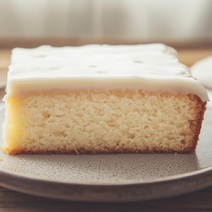 Op de afbeelding: Een vierkant stuk gele cake met witte glazuur op een grijze plaat. De cake heeft een lichte, luchtige textuur en een goudbruine korst. De plaat is rond met een licht verhoogde rand, op een houten achtergrond.