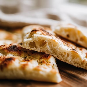 May include: Close-up of several slices of freshly baked focaccia bread. The bread has a golden-brown crust with dark, slightly charred spots. The interior is light and airy, with visible air pockets. The bread is on a wooden surface.