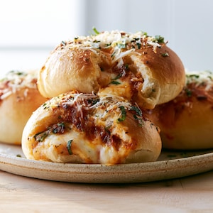 Può includere: Un gruppo di panini dorati al forno ripieni di formaggio bianco fuso e salsa di pomodoro rossa. La parte superiore è guarnita con formaggio grattugiato e pezzetti di erbe verdi, mostrando una consistenza soffice e un aspetto caldo e saporito.