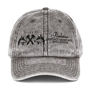 Puede incluir: Gorra de béisbol gris con un acabado desgastado. La parte delantera presenta el texto "Predation, Instinct Sharpened Into Action" y un gráfico de hachas cruzadas y cabezas de animales estilizadas. La gorra tiene una visera curva y una correa ajustable.