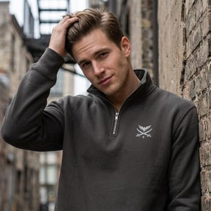 Peut inclure: Un sweat-shirt gris anthracite à fermeture éclair quart, doté d'un petit motif brodé blanc sur la poitrine gauche. Le motif représente un oiseau stylisé aux ailes déployées au-dessus de deux épées croisées et d'une petite étoile à quatre branches.