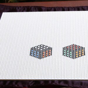 May include: White pegboard with two hexagonal designs made of colourful pegs. The pegboard has a textured surface and is set against a dark purple background. The designs feature blue, red, green, and yellow pegs.