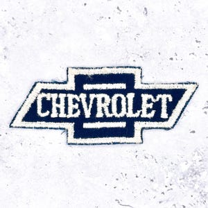 May include: A navy blue embroidered patch featuring the Chevrolet logo. The logo is a stylized bow tie shape with the word "CHEVROLET" in white capital letters. The patch has a white border and is set against a textured, light grey background.