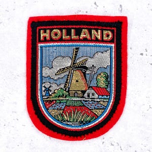 May include: Embroidered patch with the word "HOLLAND" in gold lettering. The patch features a windmill, a house with a red roof, and fields. The patch has a red border with black trim.