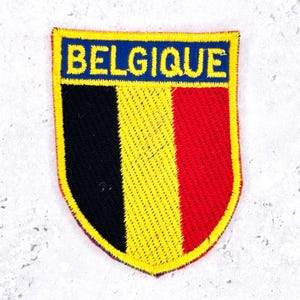 Op de afbeelding: Een schildvormige geborduurde patch met de vlag van België met verticale zwarte, gele en rode banen. Het woord BELGIQUE is in gele hoofdletters geborduurd op een blauwe horizontale balk aan de bovenzijde, geheel afgezet met een gele rand.
