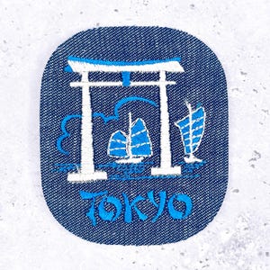 May include: Embroidered patch with a blue denim background. The design features a white Torii gate, stylized sailboats, and the word "TOKYO" in blue script. The patch has an oval shape.