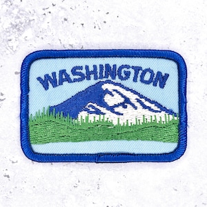 May include: A rectangular embroidered patch with a light blue background and a dark blue border. The patch features a green forest at the base, a white snow-capped mountain in the centre, and the word WASHINGTON embroidered in dark blue text.