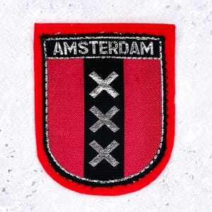 Peut inclure: Un écusson en tissu rouge de forme rectangulaire avec des coins inférieurs arrondis, présentant une bande centrale verticale noire. L'écusson affiche trois symboles X argentés brodés alignés verticalement et le mot AMSTERDAM en argent.