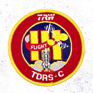 May include: Embroidered patch with a red circular border and a yellow stitched edge. The patch features a central design with a space shuttle, yellow rectangles, and the words "FLIGHT" and "TDRS-C". The text "TRW" is at the top.