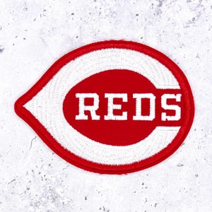 May include: An embroidered patch featuring a red and white design in the shape of a stylized letter C with a pointed left side. The word REDS is stitched in white block letters across the center of the red oval, surrounded by a white border.