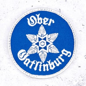 Op de afbeelding: Een ronde, blauwe patch met witte geborduurde tekst en een sneeuwvlokontwerp. De tekst luidt "Ober" bovenaan en "Gatlinburg" onderaan. De patch heeft een witte rand.