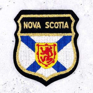 Könnte beinhalten: Gestickter Aufnäher mit dem Wappen von Nova Scotia. Das Wappen hat einen schwarzen Rand mit goldfarbenen Nähten, die Worte "NOVA SCOTIA" in Gold, einen weißen Hintergrund mit einem blauen Andreaskreuz und einen roten Löwen auf einem gelben Schild.