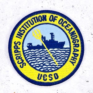 Op de afbeelding: Geborduurde patch met een marineblauwe rand en een gele buitenring. De tekst "SCRIPPS INSTITUTION OF OCEANOGRAPHY" en "UCSD" staan in het ontwerp. Een lichtblauw midden toont een schip, golven en een gele drietand.