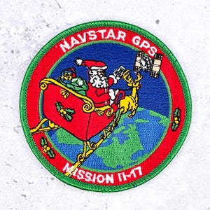 May include: A circular embroidered patch featuring Santa Claus in a red sleigh pulled by a yellow reindeer over a blue and green Earth. The text NAVSTAR GPS and MISSION 11-17 are in white on a red border. A satellite is visible in the blue sky.