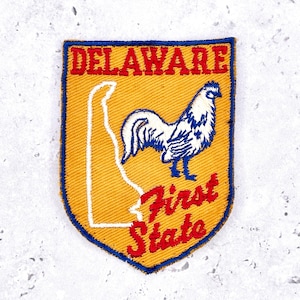 May include: Embroidered patch featuring the word "DELAWARE" in red, arched across the top. Below, a white outline of the state and a blue and white rooster. The words "First State" are in red script at the bottom. The patch has a yellow background and a blue border.