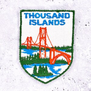 May include: Embroidered patch featuring the words "THOUSAND ISLANDS" in blue above an orange bridge design. The bridge spans blue water with green trees along the bottom edge. The patch has a white background and a green border.