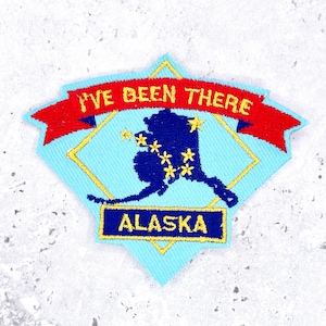 May include: A light blue embroidered patch shaped like a shield with a red banner at the top reading I'VE BEEN THERE in yellow. The center features a dark blue map of Alaska with yellow stars and a yellow rectangle at the bottom reading ALASKA.