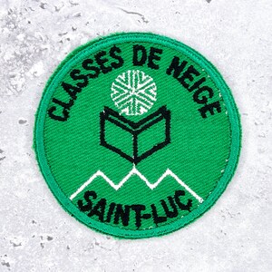 May include: A green circular patch with the text "CLASSES DE NEIGE SAINT-LUC" in black. The patch features a white snowflake, an open book, and a mountain range design.