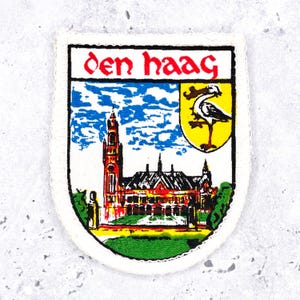 Op de afbeelding: Een schildvormige geborduurde patch met de tekst den haag in rode letters. Het ontwerp toont een gedetailleerde illustratie van een gebouw met een hoge toren onder een blauwe lucht, naast een geel wapenschild met een zwart-witte ooievaar.