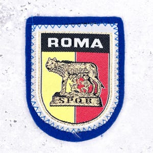 May include: A rectangular embroidered patch with a blue border featuring the word ROMA in white text on a black background. Below, a yellow and red shield displays a golden she-wolf nursing two infants above the letters SPQR in gold thread.