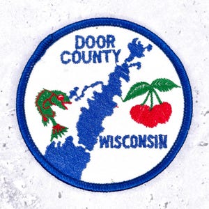 May include: A circular embroidered patch with a white background and a blue border. It features a blue map of Door County, Wisconsin, with the text DOOR COUNTY WISCONSIN in blue, a green fish with red accents, and two red cherries with green leaves.