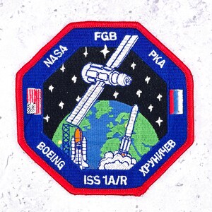 May include: Embroidered patch with a blue octagonal border and a red trim. The patch features a space station, Earth, and a rocket launching. Text includes "NASA", "BOEING", "ISS 1A/R", "FGB", "PKA", and "ХРУНИЧЕВ".