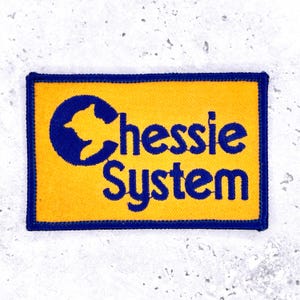 Op de afbeelding: Geborduurde patch met een gele achtergrond en een blauwe rand. De patch heeft een blauw silhouet van een vogel in een blauwe cirkel, met de woorden "Chessie System" in blauw.