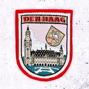May include: Embroidered patch with a red border featuring the text "DEN HAAG" at the top. The patch displays a detailed illustration of a building and a coat of arms with a stork. The background is pink.