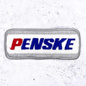 May include: Embroidered patch with the word "PENSKE" in red and blue letters on a white background. The patch has a gray border and a rectangular shape.