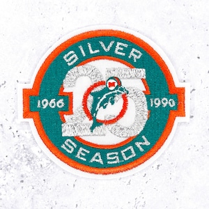 May include: Embroidered patch with a white background. The patch features a teal and orange circular design with the words "SILVER SEASON" and "1966 1990". A dolphin logo is in the center. The patch is a commemorative item.