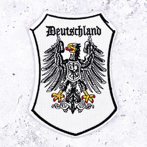 May include: A shield-shaped embroidered patch featuring the word Deutschland in black gothic lettering above a black eagle with yellow talons and beak, a red tongue, and a smaller central shield, all outlined in a thick black border.