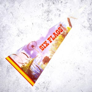 Könnte beinhalten: Eine dreieckige Wimpelfahne mit den Worten "SIX FLAGS" in Rot. Die Fahne zeigt eine farbenfrohe Vergnügungspark-Szene mit Achterbahnen, einem Riesenrad und anderen Attraktionen. Die Fahne hat einen gelb-roten, genähten Rand.