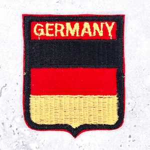 May include: Embroidered patch featuring the German flag design. The patch has a rectangular shape with a black stripe, a red stripe, and a yellow stripe at the bottom. The word "GERMANY" is embroidered in gold letters at the top.