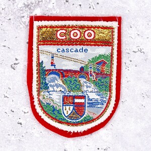 Può includere: Toppa rettangolare con bordo rosso e design ricamato dettagliato. Il design presenta un ponte, cascate e la parola "cascade". La parte superiore della toppa presenta la scritta "COO" in lettere dorate.