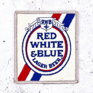 May include: Square patch with a white background and a red, white, and blue design. The patch features the text "RED WHITE & BLUE LAGER BEER" in blue, with a red and blue stripe design. The patch has a stitched border.
