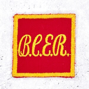 May include: A square-shaped embroidered patch featuring a vibrant red background with a thick yellow border. The word "BEER" is stitched in the center in a bold, yellow, cursive-style font, creating a clear contrast against the red fabric.