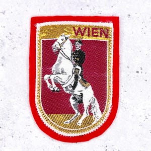 May include: A red and gold embroidered patch featuring a white horse rearing up, ridden by a person in a black coat. The word "WIEN" is at the top. The patch has a red border and a gold trim.