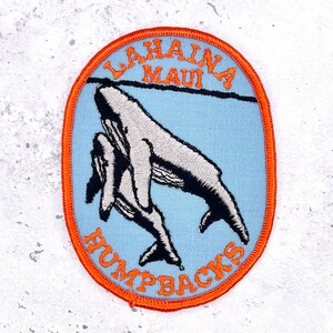 May include: An oval embroidered patch with a light blue background and a bright orange border. It features a black and white illustration of two humpback whales swimming. The text LAHAINA MAUI is embroidered in orange at the top, and HUMPBACKS is at the bottom.
