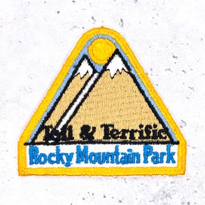 May include: Embroidered patch in a triangular shape with a yellow border. The design features a mountain range with a sun, the text "Chill & Terrific" and "Rocky Mountain Park" in blue.