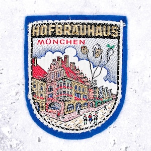 Può includere: Una toppa ricamata blu con il testo HOFBRAUHAUS MÜNCHEN in filo dorato e rosso. Il disegno raffigura un edificio a più piani con un tetto rosso, un cielo bianco con nuvole e tre boccali di birra fluttuanti. La toppa ha un bordo bianco cucito.