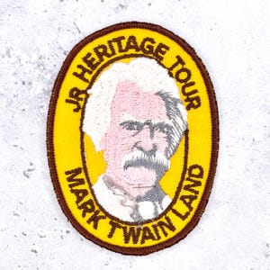 May include: An oval embroidered patch with a yellow background and a brown border. It features a portrait of Mark Twain in the center with white hair and a mustache. The text "JR HERITAGE TOUR" and "MARK TWAIN LAND" is stitched in brown.