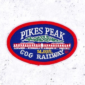 May include: An oval embroidered patch with a red border and a blue background. It features white text reading PIKES PEAK COG RAILWAY, a green mountain graphic, a white train illustration, and yellow text stating 14,110 ft.