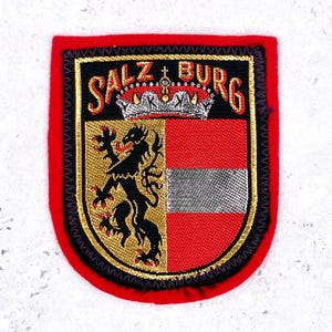 May include: A shield-shaped embroidered patch with a red border. The center features a black lion on a gold background next to a red and silver horizontal striped field. Above this, a silver crown sits below the text SALZ BURG in red.
