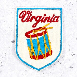May include: A shield-shaped embroidered patch with an off-white background and a blue border. The text Virginia is stitched in red script above a graphic of a blue drum with red rims and yellow accents, including two crossed drumsticks.