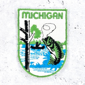 Może przedstawiać: Haftowana naszywka z zieloną obwódką i napisem "MICHIGAN" u góry. Naszywka przedstawia scenę wędkarską z czarnym drzewem, łodzią i wyskakującą rybą. Ryba jest zielono-czarna, a woda niebieska.