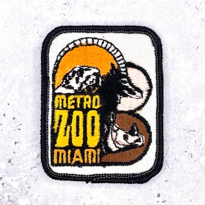 Może przedstawiać: Haftowana naszywka z napisem "Metro Zoo Miami" w kolorze żółtym oraz czarno-biało-brązowym wzorem zwierzęcym. Naszywka ma czarną obwódkę i białe tło, z pomarańczowym kształtem przypominającym słońce.
